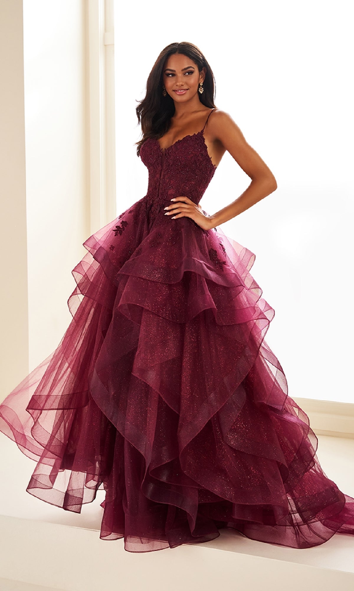 A woman stands indoors before a bright window, wearing the Ellie Wilde EW36203 Glitter Tulle Prom Ball Gown—sleeveless, dark red, with layered tulle skirt, an embroidered corset bodice, and adjustable lace-up back.
